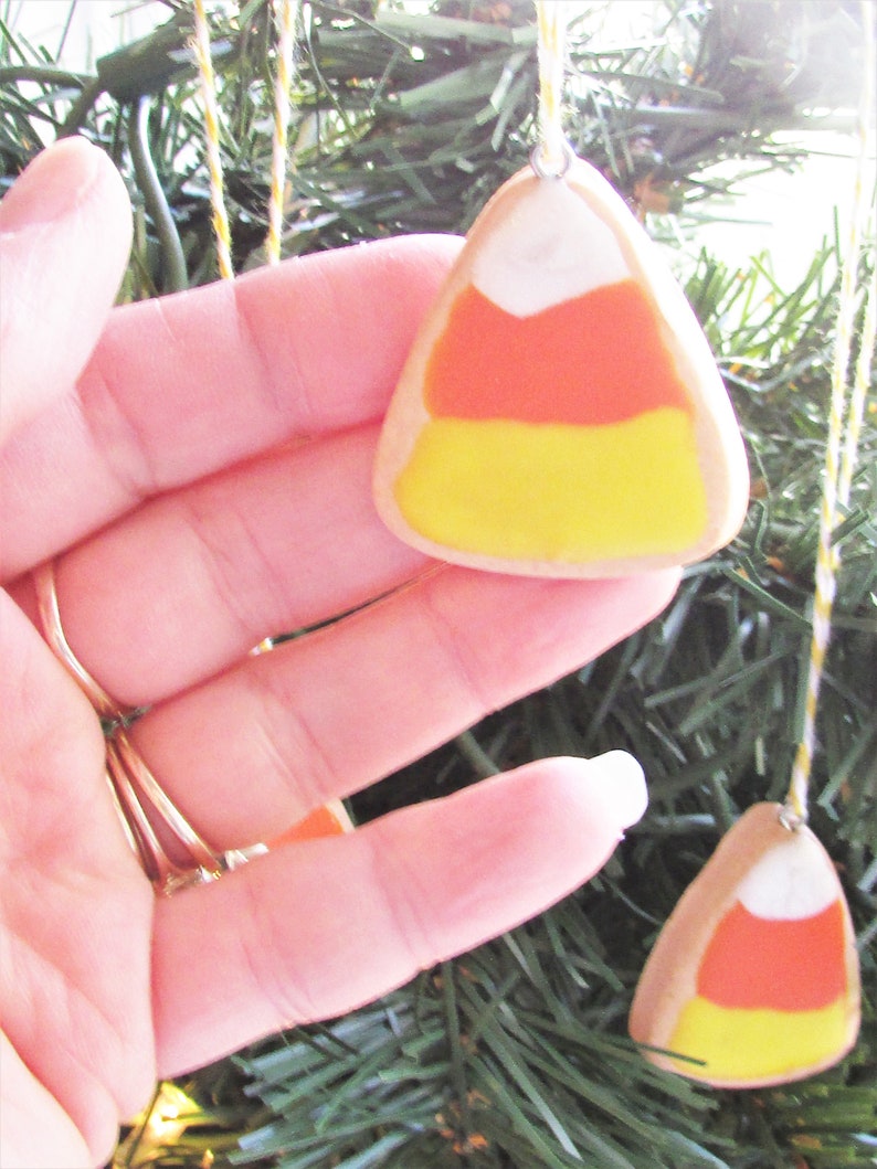 Puede incluir: Una mano sostiene un adorno de ma&iacute;z dulce amarillo, naranja y blanco. El adorno est&aacute; colgado de una cuerda en un &aacute;rbol de Navidad artificial verde.