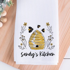 May include: White kitchen towel with a yellow and black beehive design and the text "Sandy's Kitchen".