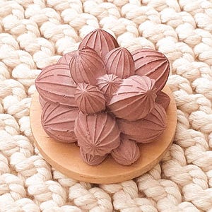 May include: A cluster of small, textured, reddish-brown decorative spheres sits atop a round, light-brown wooden base. The spheres have a ribbed surface. The base is placed on a cream-colored, chunky knit surface.