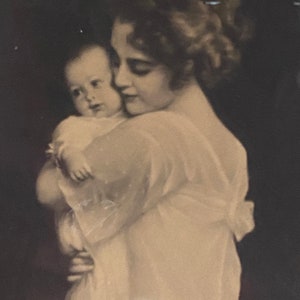 May include: A black and white photograph of a woman holding a baby in her arms. The woman is wearing a white dress and the baby is wearing a white dress. The woman is looking down at the baby and the baby is looking up at the woman.