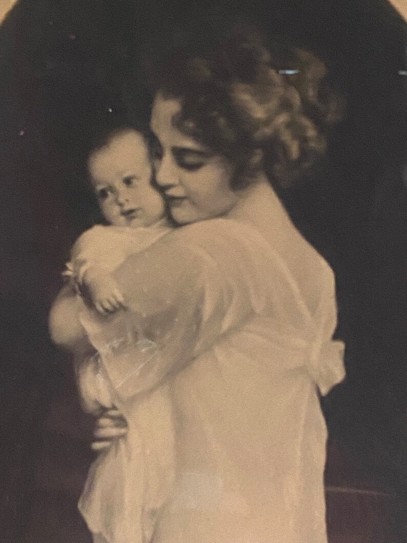 May include: A black and white photograph of a woman holding a baby in her arms. The woman is wearing a white dress and the baby is wearing a white dress. The woman is looking down at the baby and the baby is looking up at the woman.