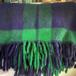 Puede incluir: Una manta de lana a cuadros en verde y azul marino con flecos. La manta está hecha de un material suave y peludo.
