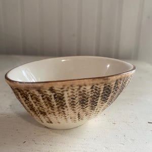 May include: A small, oval-shaped ceramic bowl with a smooth, off-white interior. The exterior features a textured, brown, woven-like pattern that wraps around the sides, finished with a dark brown rim along the top edge of the bowl.