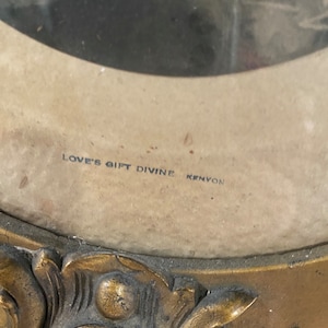 May include: A gold-colored ornate picture frame with a faded photograph inside. The frame has a decorative design and the text "LOVE'S GIFT DIVINE KENYON" is printed on the bottom.