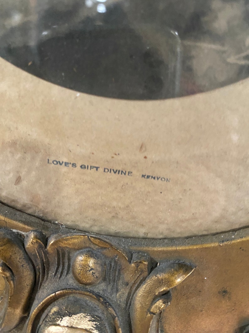May include: A gold-colored ornate picture frame with a faded photograph inside. The frame has a decorative design and the text "LOVE'S GIFT DIVINE KENYON" is printed on the bottom.