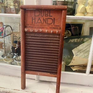 Op de afbeelding: Een vintage houten wasbord met de tekst "DUBL HANDI" in zwarte letters. Het wasbord heeft een roodbruin frame en een reeks horizontale ribbels om kleding te schrobben. Haken zijn aanwezig aan de bovenkant. Het wasbord is een antiek wasgereedschap.