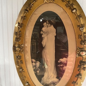 May include: An oval-shaped picture frame with a gold finish and ornate detailing. The frame contains a black and white photograph of a woman holding a child.