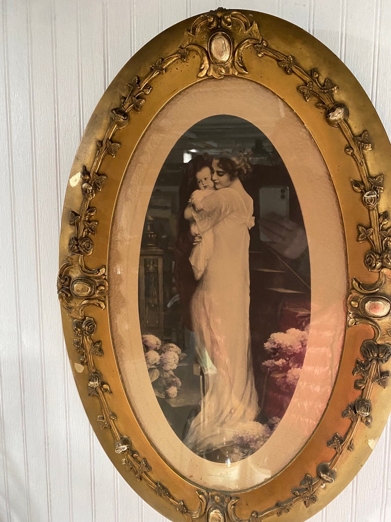 May include: An oval-shaped picture frame with a gold finish and ornate detailing. The frame contains a black and white photograph of a woman holding a child.