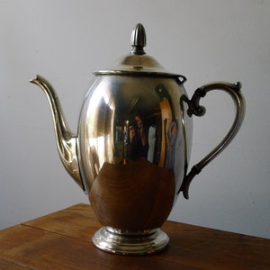 Puede incluir: Una tetera de plata con una superficie brillante y reflectante. La tetera tiene un cuerpo redondeado, un pico, una asa y una tapa con un pomo.