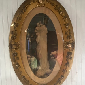 May include: An oval-shaped gold picture frame with ornate details and a faded image of a woman holding a child. The image is set against a dark background with flowers in the foreground.