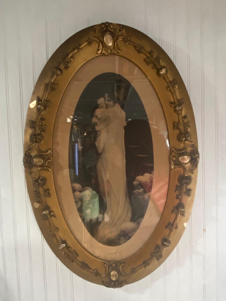 May include: An oval-shaped gold picture frame with ornate details and a faded image of a woman holding a child. The image is set against a dark background with flowers in the foreground.