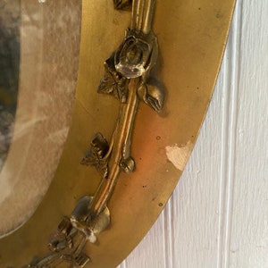 May include: A gold oval mirror with a decorative rose design around the frame. The mirror is slightly tarnished and has some minor scratches.