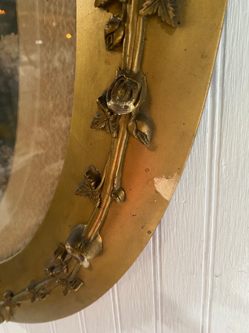 May include: A gold oval mirror with a decorative rose design around the frame. The mirror is slightly tarnished and has some minor scratches.