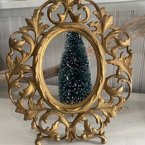 Puede incluir: Un marco de fotos de metal dorado con un diseño ornamentado. El marco tiene una parte superior decorativa y un soporte en la parte inferior. El marco está vacío.