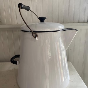 Op de afbeelding: Witte emaille koffiepot met een zwarte handgreep en dekselknop. De pot heeft een tuit en een draadhandvat. De rand is zwart omlijnd. Een vintage keukenartikel.