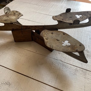 May include: A vintage pair of ice skates with a wooden base and metal blades. The blades have a cloverleaf design and are attached to the base with a metal hinge.
