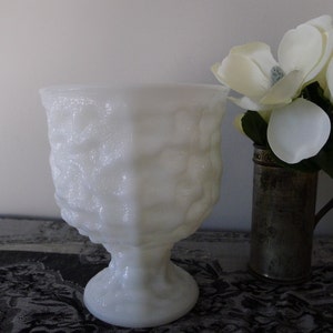 Könnte beinhalten: Weiße Milchglasvase mit strukturierter Oberfläche. Die Vase hat einen breiten, runden Boden und einen hohen, zylindrischen Körper. Die Vase steht auf einer schwarz-weiß gemusterten Oberfläche.