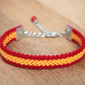 Op de afbeelding: Een rood en geel geweven armband met een zilveren sluiting. De armband is gemaakt van dunne koorden en heeft een eenvoudig ontwerp.