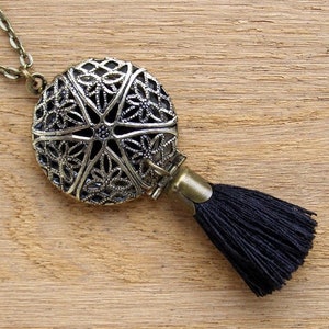Puede incluir: Un collar con un medallón plateado y una borla negra. El medallón tiene un diseño de filigrana y está abierto.