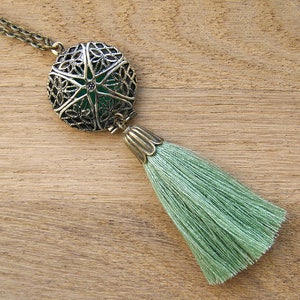 Puede incluir: Un collar con un colgante redondo de bronce adornado. El colgante tiene un diseño de filigrana y una piedra verde en el centro.