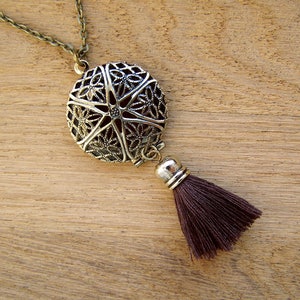 Puede incluir: Un collar plateado con un colgante redondo de filigrana y un borla marrón. El colgante tiene un diseño floral delicado.