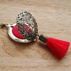 Puede incluir: Un collar con un medallón de oro, un interior de fieltro rojo y una borla roja. El medallón está abierto y tiene un delicado diseño de filigrana.