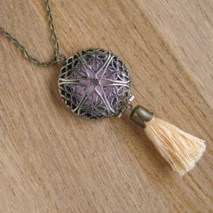 Puede incluir: Un collar de bronce con un colgante redondo de filigrana y un interior rosa claro. El colgante tiene una borla de tela de color crema.