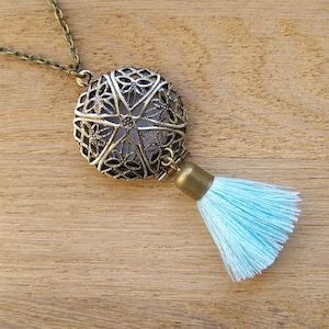 Puede incluir: Un collar de bronce con un colgante redondo de filigrana y un borla azul claro. El colgante tiene una tapa con bisagras y está diseñado para contener objetos pequeños.