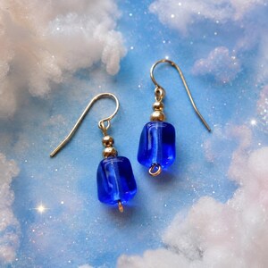 Può includere: Un paio di orecchini pendenti con perline di vetro blu cobalto di forma rettangolare. Ogni orecchino è accentuato da una piccola perlina di metallo dorato sopra il vetro e pende da un semplice gancio color oro per lobi forati.