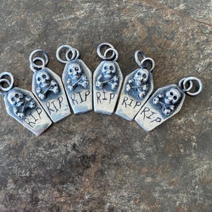Könnte beinhalten: Sechs silberfarbene Anhänger in Sargform mit Totenkopf und gekreuzten Knochen sowie der Gravur "RIP". Die Anhänger haben eine Schlaufe zum Befestigen. Der Hintergrund ist eine strukturierte, dunkle Oberfläche. Ideal für Halloween.