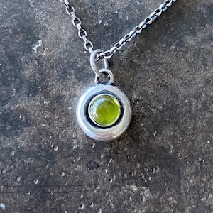 Op de afbeelding: Een zilveren ketting met een ronde hanger met een groene edelsteen.
