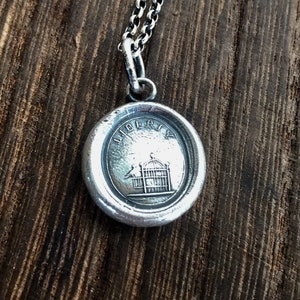 以下が含まれることがあります： 鳥かごの中に2羽の鳥が入った「LIBERTY」という文字が刻まれた丸いペンダントが付いたシルバーネックレス