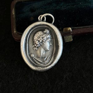 Puede incluir: Pendiente de plata con un perfil detallado de una mujer con una corona de laurel, en un marco ovalado.