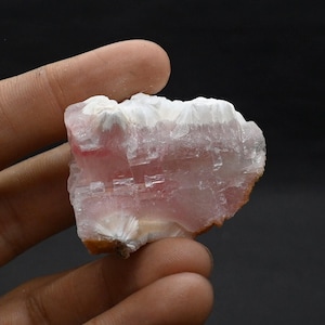 May include: A raw, natural mineral specimen with a layered structure. The stone features a mix of translucent pink and white hues, with some areas appearing crystalline. The edges have a rough, uneven texture, and the overall shape is irregular.