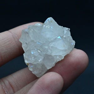 Può includere: Un gruppo di cristalli di quarzo trasparente con lampi iridescenti di colore. I cristalli hanno una consistenza ruvida e naturale e sono tenuti in mano su uno sfondo scuro. La formazione di quarzo ha una forma appuntita e irregolare.