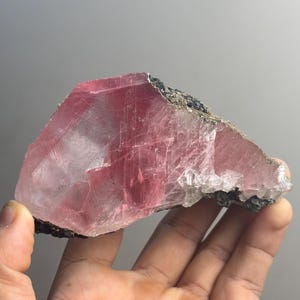 以下が含まれることがあります： 半透明で鮮やかなピンク色の結晶構造を持つ未加工の鉱物標本。内部には目に見える亀裂やファセットがあり、底面と側面に沿って暗く粗い質感の母岩が付着しています。