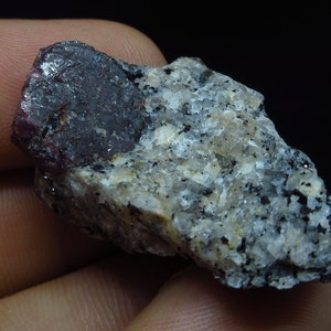 May include: A rough, grey rock with a large, dark red ruby crystal embedded in it.
