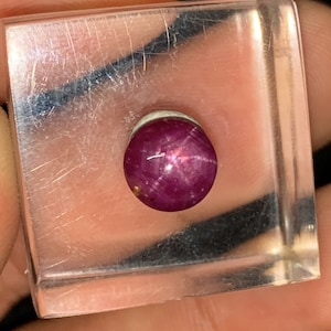Puede incluir: Una piedra preciosa de rubí estrella rojo oscuro con una estrella de seis puntas en el centro, colocada en una vitrina cuadrada transparente.