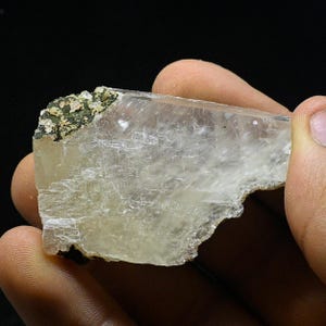 Könnte beinhalten: Ein durchscheinender, hellbeiger, natürlich geformter Kristall mit einer rauen, unregelmäßigen Form. Der Kristall hat an einem Ende eine dunkelgrüne und braune Mineraleinschlüsse.