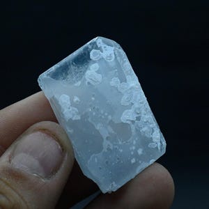 Puede incluir: Un cristal transparente y translúcido con forma rectangular y una superficie rugosa y texturizada. El cristal tiene una apariencia blanca y nublada.