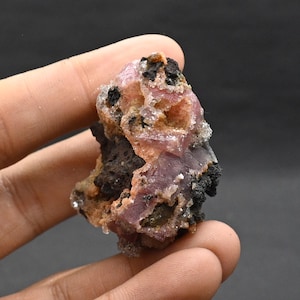 Könnte beinhalten: Nahaufnahme einer Mineralprobe mit Farbmix. Die Probe hat einen rosa-violetten Farbton mit schwarzen und beigen Bereichen. Das Mineral scheint eine kristalline Struktur zu haben und wird in einer Hand vor dunklem Hintergrund gehalten.