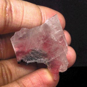 Könnte beinhalten: Ein durchscheinender rosa und grauer Kristall mit einer geschichteten Struktur. Der Kristall hat eine raue, unebene Oberfläche und ist teilweise von einem dunkelgrauen Mineral bedeckt.