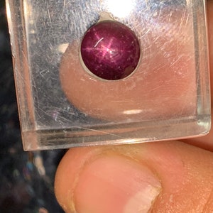 May include: A deep red star ruby gemstone with a six-pointed star in the center, set in a clear square display case.