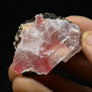 Könnte beinhalten: Ein rosa und weißer Kristall mit einer rauen, schwarzen Kante. Der Kristall hat eine durchscheinende Erscheinung und besteht aus vielen kleinen, flachen Facetten.