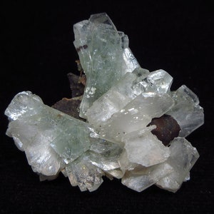 Op de afbeelding: Een cluster van doorschijnende, lichtgroene en witte kristalformaties. De kristallen hebben een gelaagde, geometrische structuur, met sommige gebieden die meer ondoorzichtig lijken. Een donkerbruine basis ondersteunt de kristallen tegen een zwarte achtergrond.