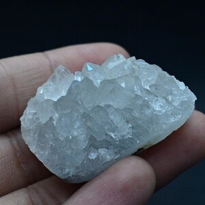 Può includere: Un gruppo di cristalli traslucidi blu chiaro. I cristalli hanno una superficie ruvida e strutturata e sono tenuti in una mano su uno sfondo scuro. I cristalli hanno una forma naturale e organica.