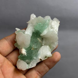 Könnte beinhalten: Ein rohes Mineralexemplar mit einer Ansammlung durchscheinender, blassgrüner Kristallstrukturen, die mit undurchsichtigen, weißen, faserigen und blockartigen Mineralformationen durchsetzt sind und eine texturierte geologische Formation bilden.