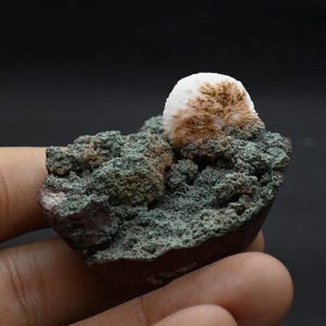 以下が含まれることがあります： 鉱物標本のクローズアップ。茶色のアクセントが付いた白い球状の形成が、緑と茶色のテクスチャベースの上にあります。鉱物は暗い背景にあります。