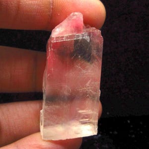 Pode incluir: Um cristal transparente, rosa e branco com forma retangular. O cristal tem um leve tom rosado e é translúcido.