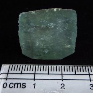 Może przedstawiać: Przezroczysty, jasnozielony kryształ o szorstkiej, fakturowanej powierzchni. Kryształ ma około 2 cm długości i leży na białej linijce.